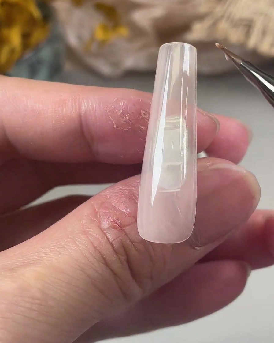 Un pennello di precisione stende uno strato sottile di gel base trasparente su una tip lunga e squadrata color nude.