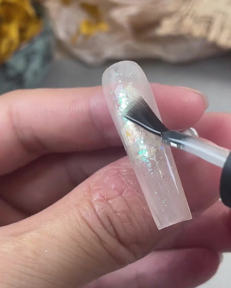 Un pennello applica un generoso strato di gel trasparente rinforzante su ununghia decorata con glitter iridescenti.