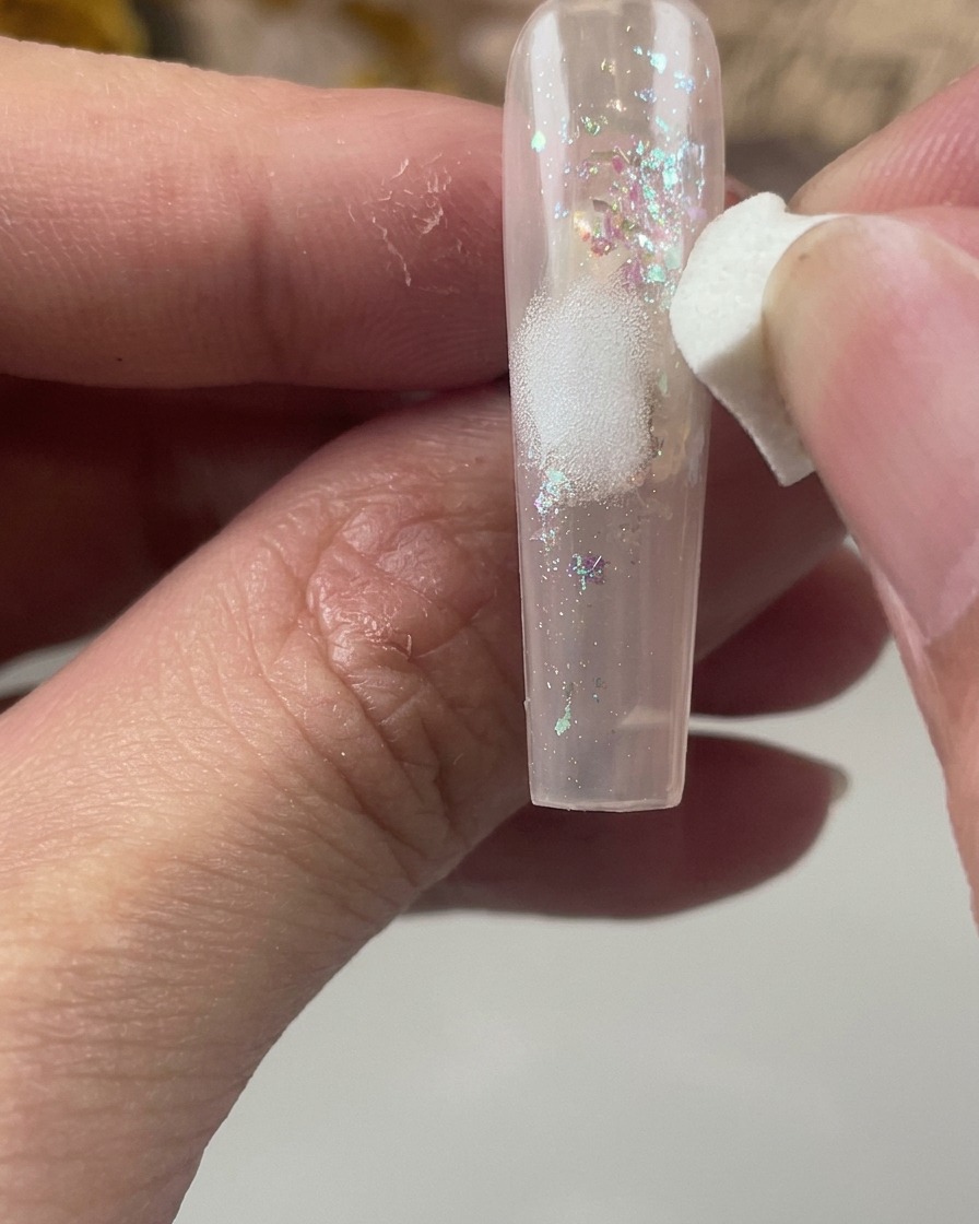 Una spugnetta cosmetica bianca tampona delicatamente una macchia di colore bianco al centro di ununghia incapsulata con glitter.
