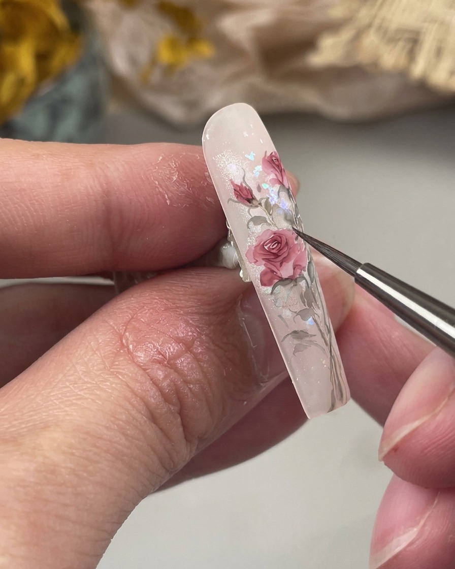 Uninquadratura ravvicinata mostra un pennellino fine che dipinge piccole foglie verdi affusolate alla base della nail art floreale color rosa sullunghia.