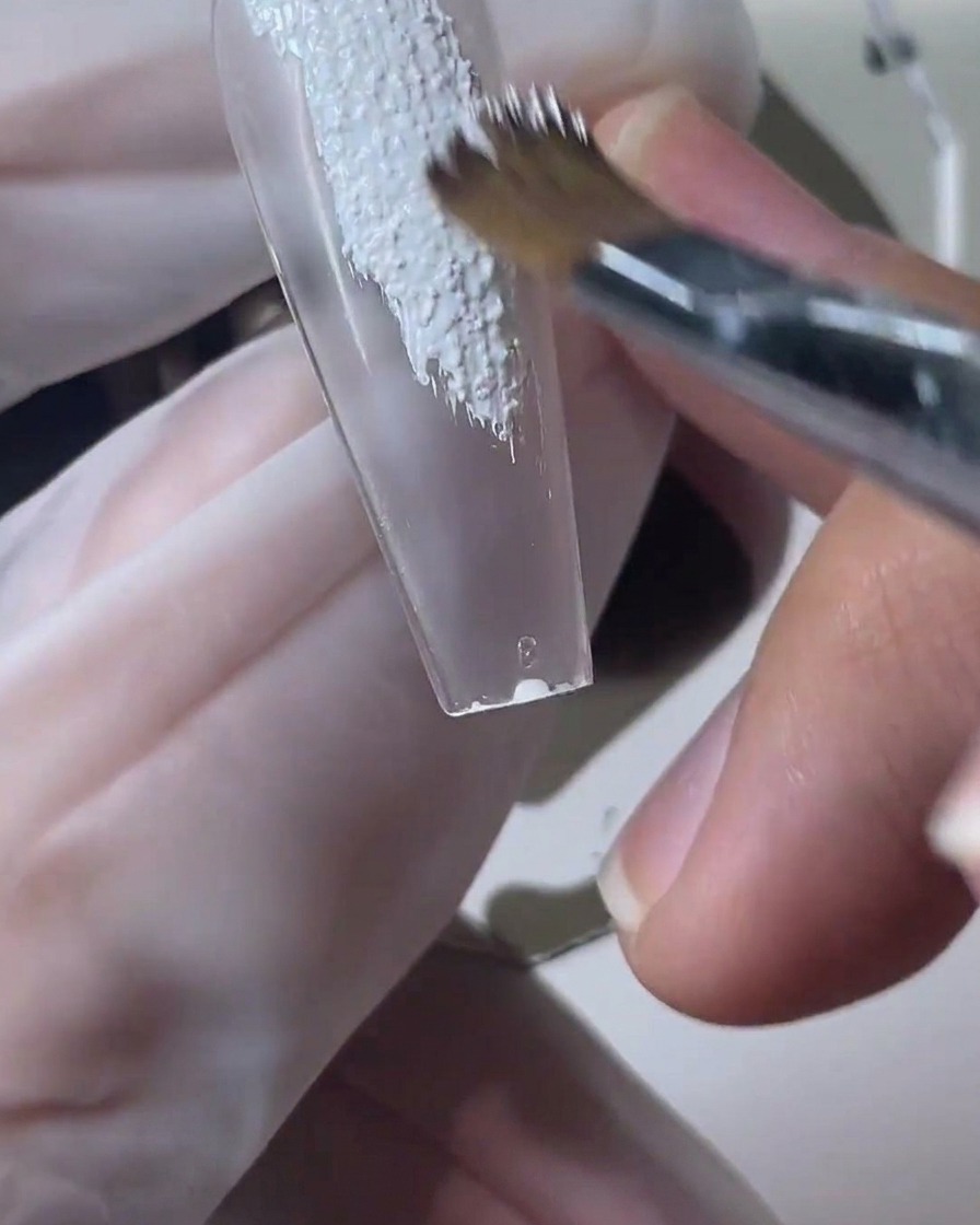 Un pennello per unghie applica gel bianco effetto sabbia su una tip trasparente.