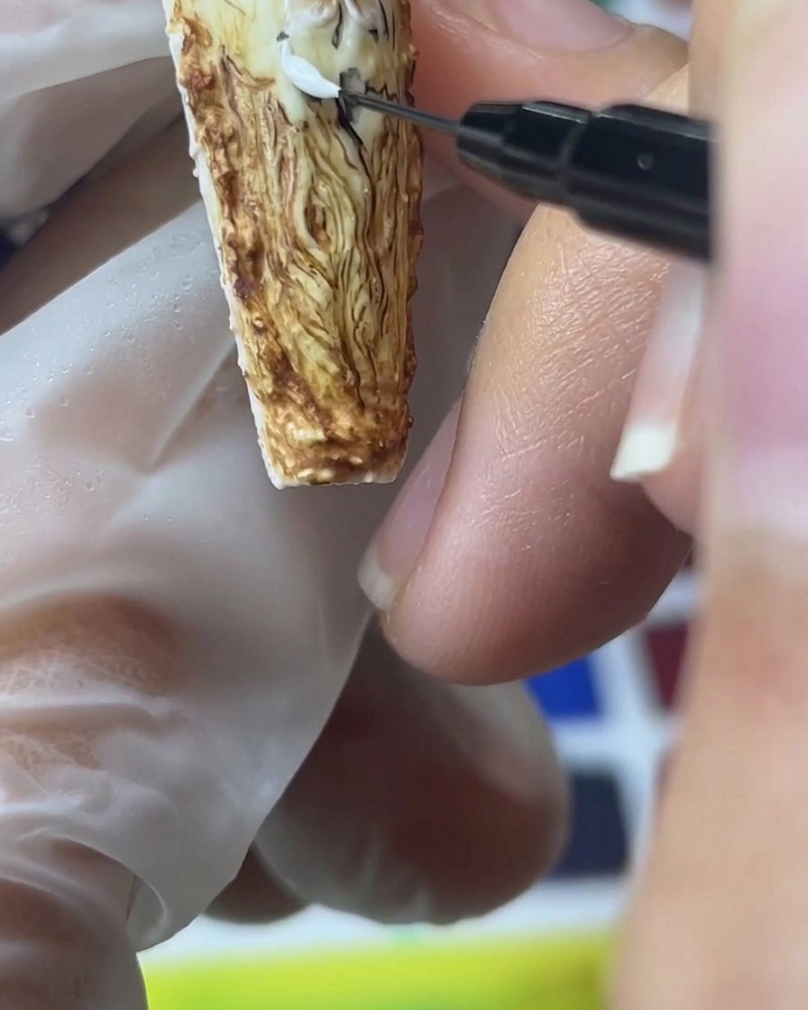 Vista ravvicinata di un pennello di precisione che applica gel sabbia bianco per creare petali di rosa 3D testurizzati su uno sfondo marrone.