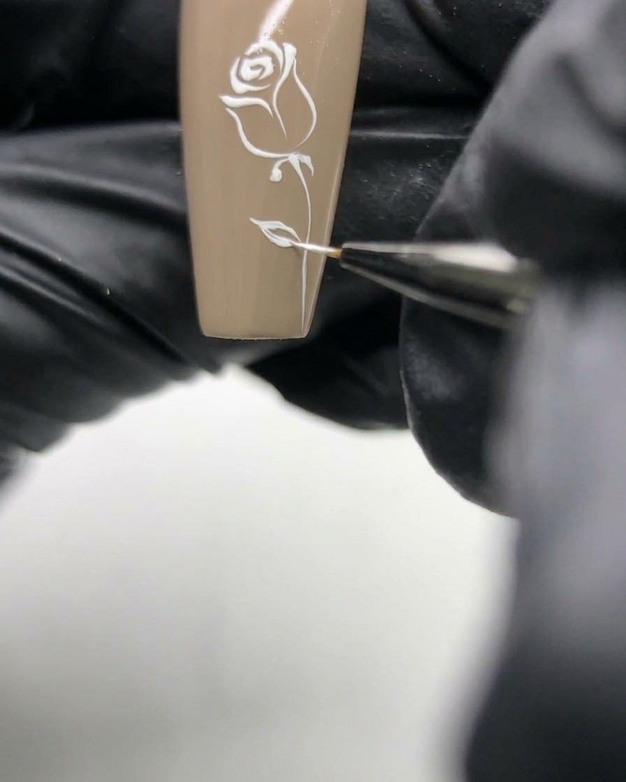 Makroaufnahme einer Nageldesignerin, die die feinen weißen Linien der Rose auf einer neutralen Basis nachzieht.