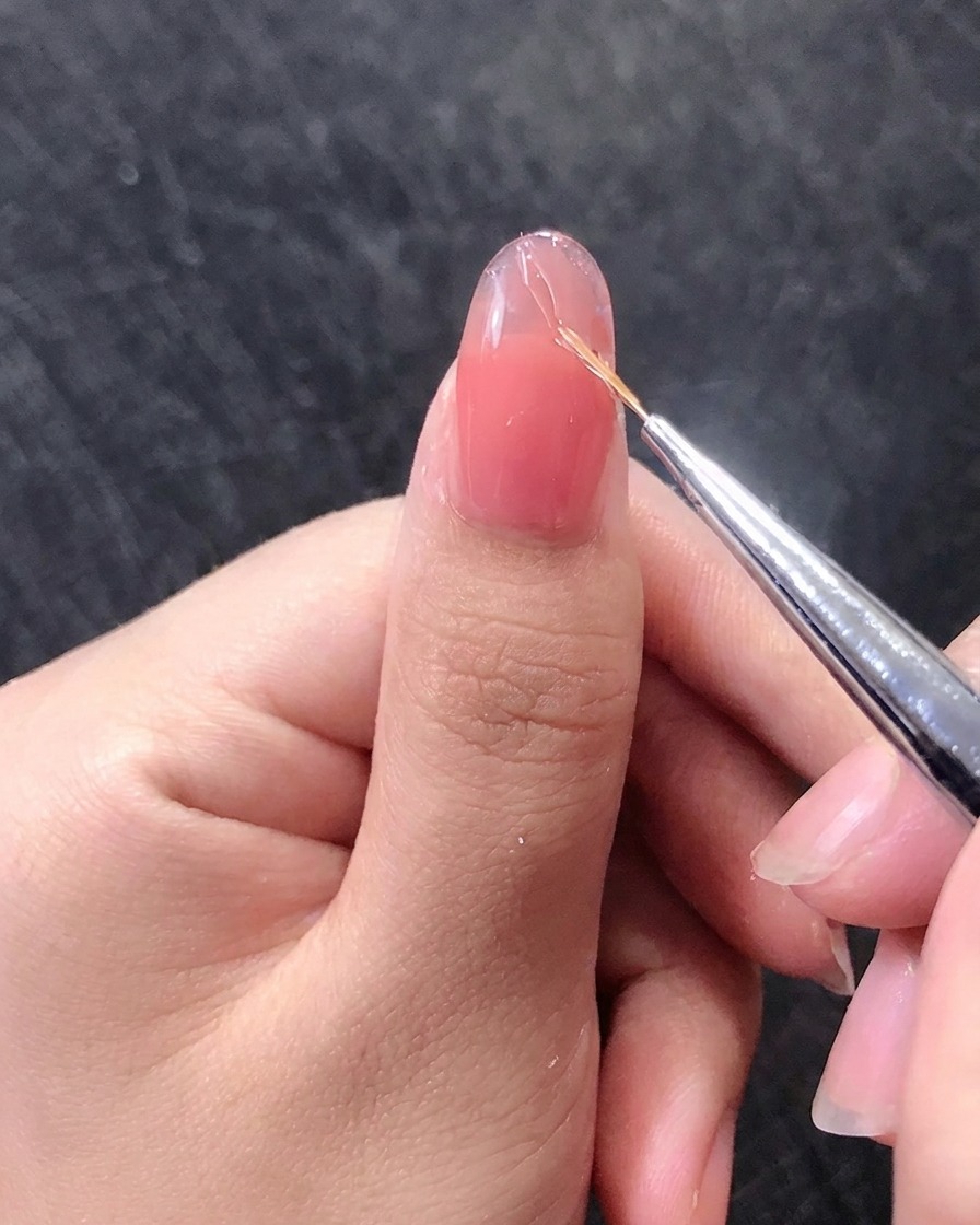 Primo piano di un pennellino che disegna linee sottili dorate su ununghia in gel rosa.