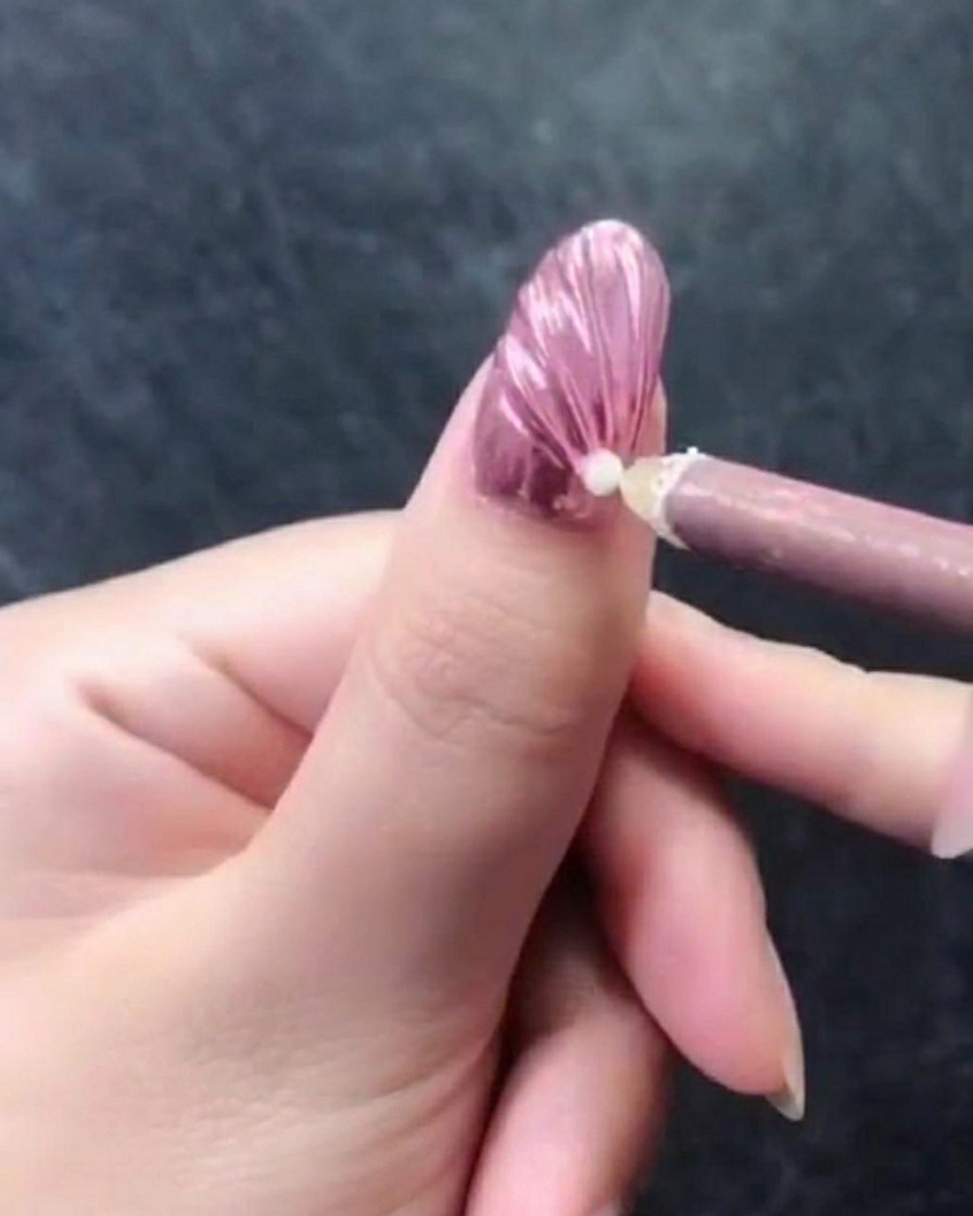 Primo piano di una mano con ununghia rosa metallizzata in rilievo, mentre una matita in cera posiziona una singola perla bianca vicino alle cuticole.