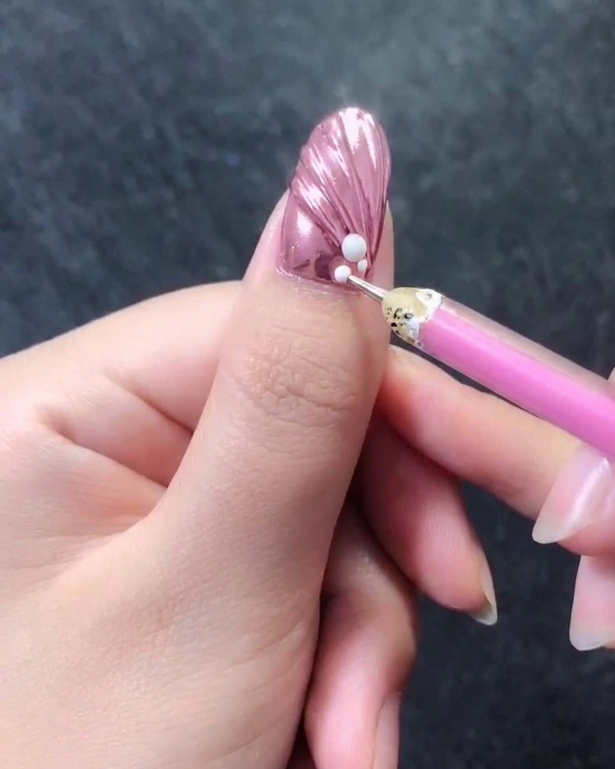 Primo piano di ununghia rosa metallizzata in rilievo con una matita in cera che aggiunge minuscole perline bianche accanto a una perla più grande.