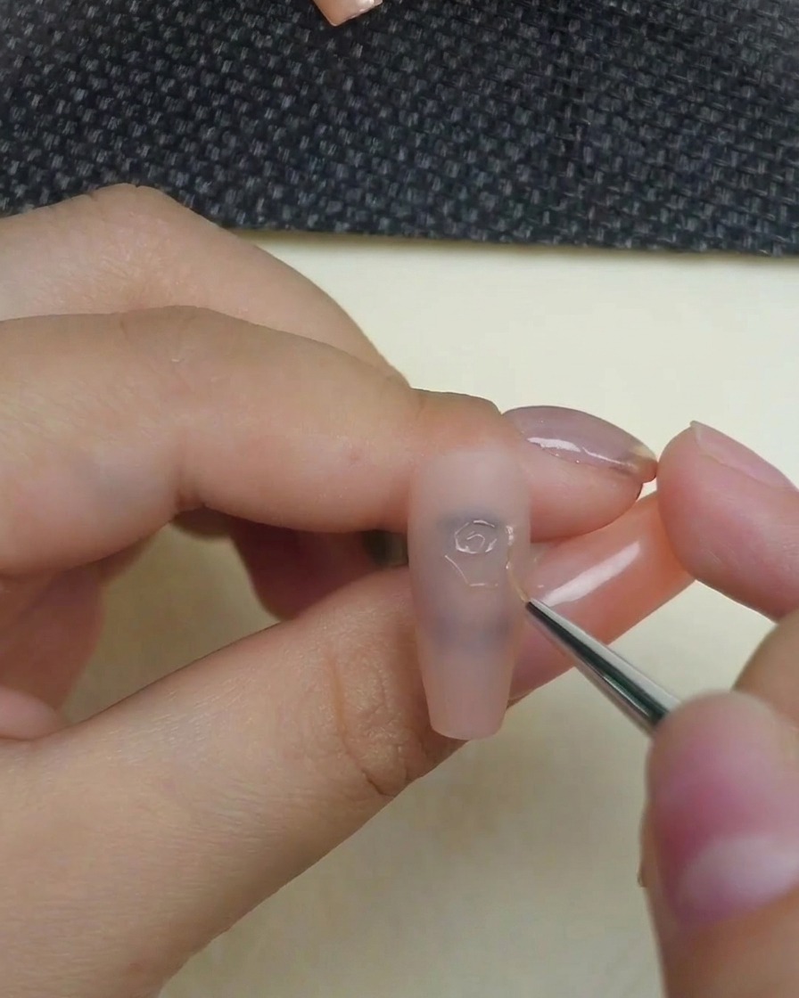 Primo piano di una mano che utilizza un pennello a punta fine con gel per scultura 3D trasparente per disegnare le spirali centrali di una rosa su ununghia color carne opaca.