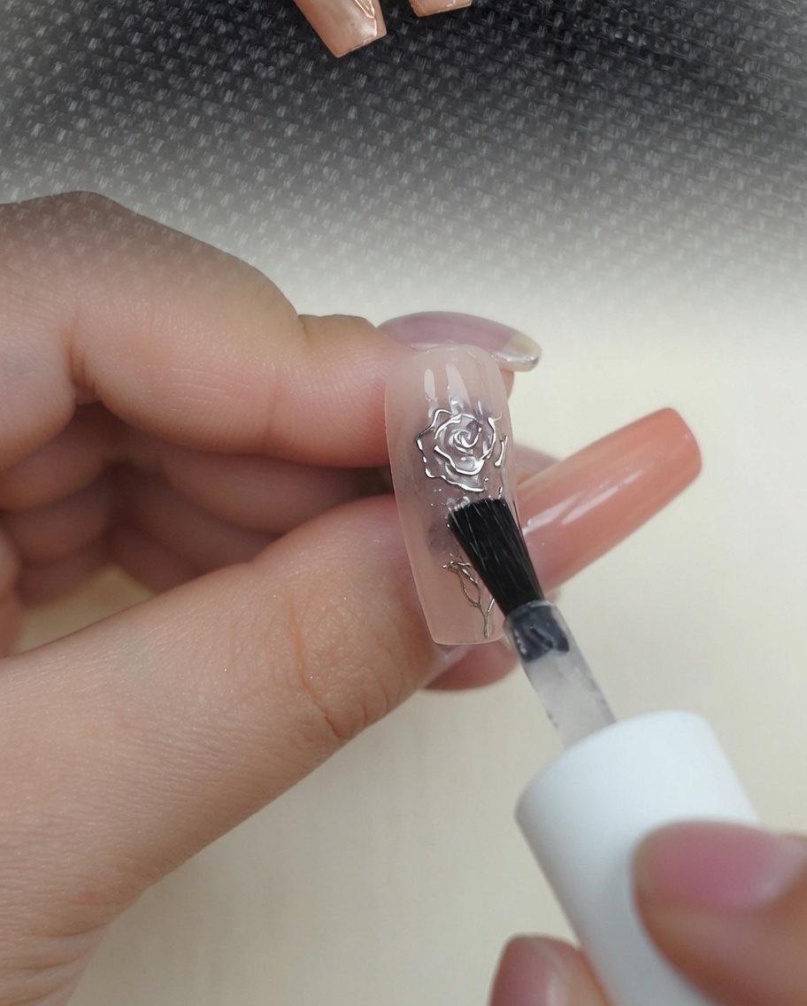 Un pennello applica un top coat lucido e trasparente sopra una rosa 3D metallizzata su ununghia color carne.