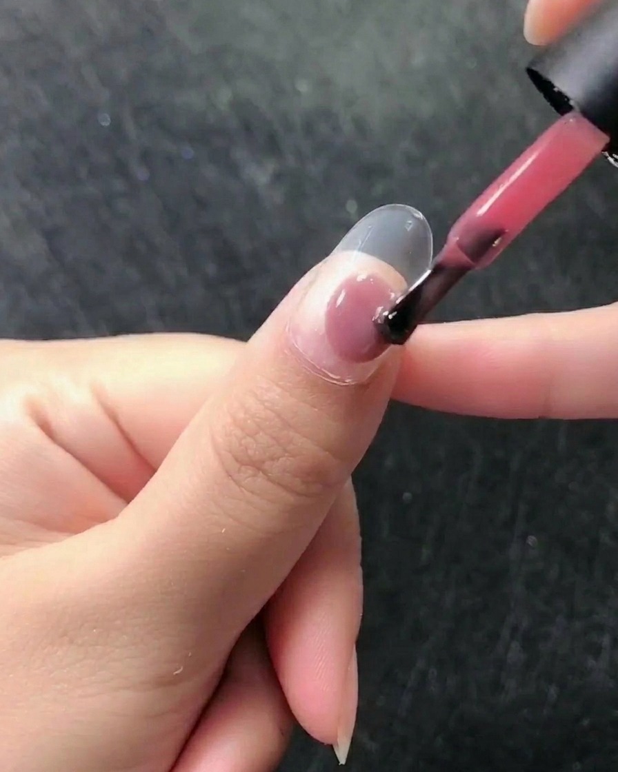 Primo piano di un pennello che applica uno smalto gel rosa velato su ununghia naturale e preparata.