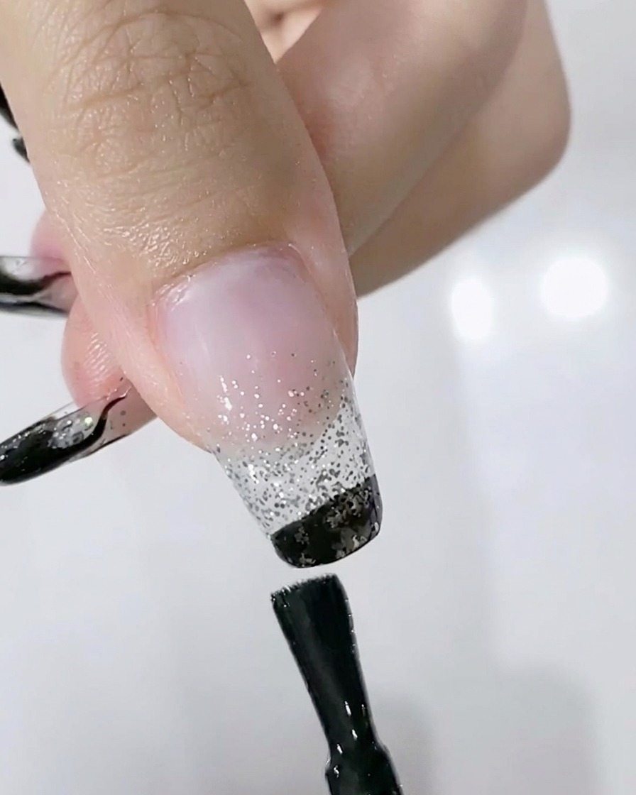 Un pennello che applica smalto gel nero sul bordo libero di ununghia sopra uno sfumato glitter argento.