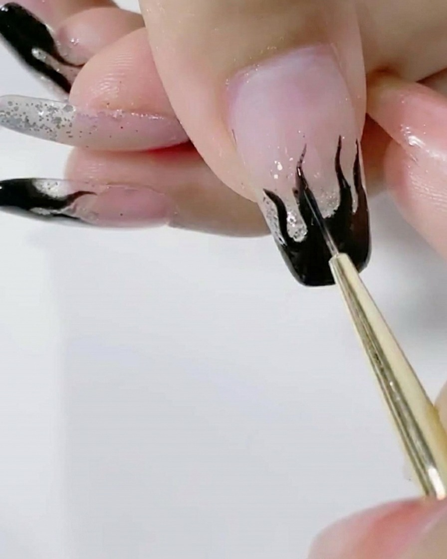 Un pennello liner che applica un secondo strato di gel nero saturo sulla decorazione a fiamma su ununghia glitter.