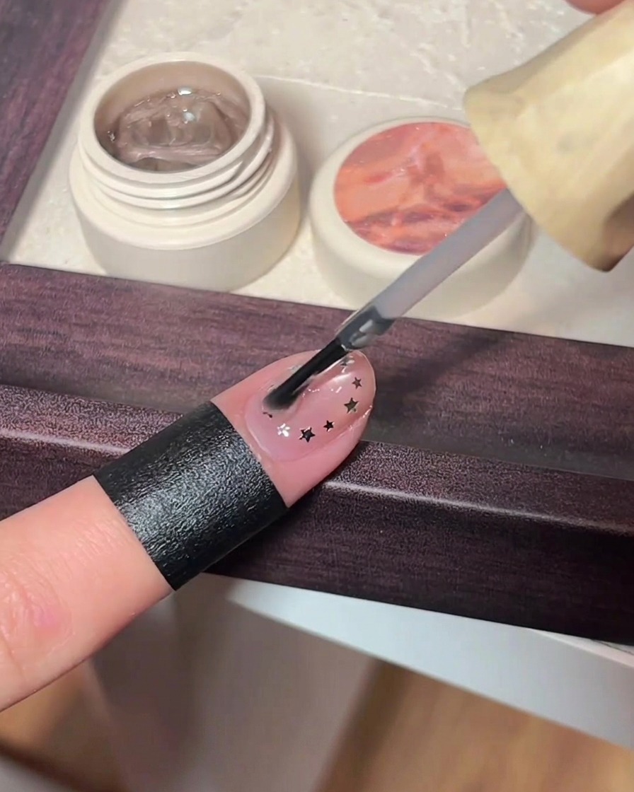 Pennello che applica uno strato generoso di top coat trasparente su unghie con glitter a stella.