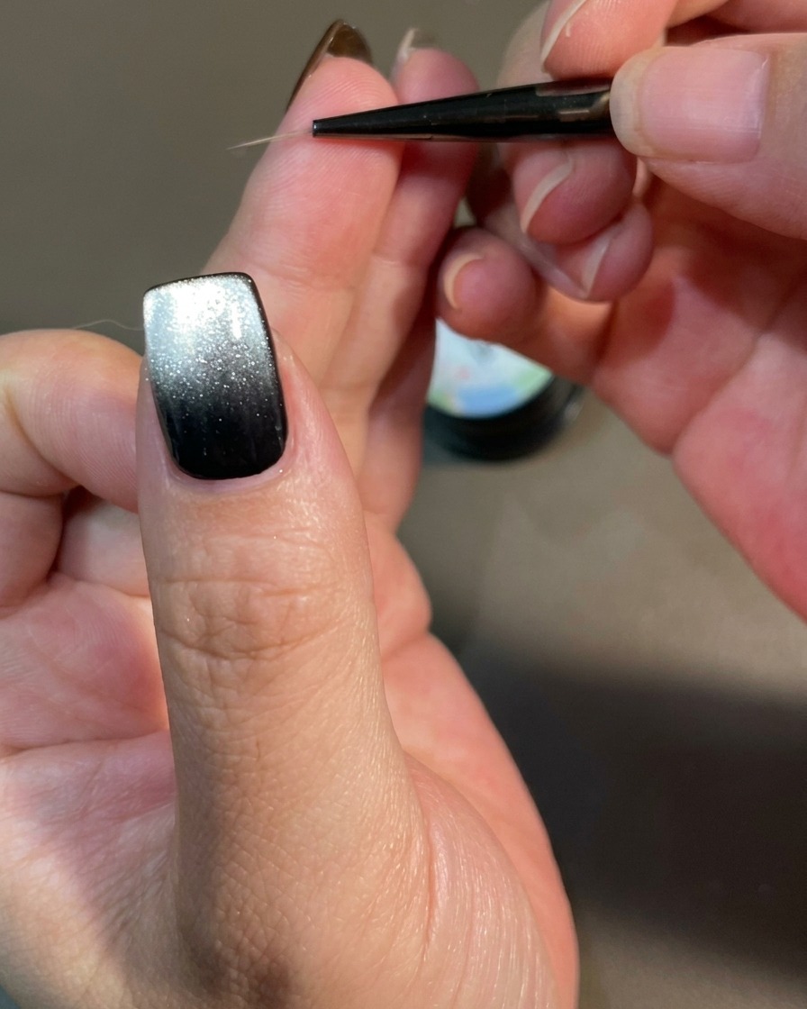 Un primo piano di ununghia decorata con un pennello liner sottile che crea linee curve simili a fiamme su una base di gel magnetico nero.