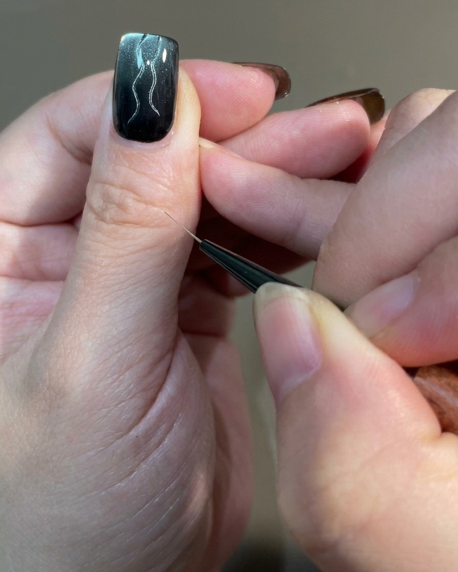 Un primo piano di ununghia che mostra linee curve di diverse lunghezze disegnate con un pennello liner su una base di gel magnetico.