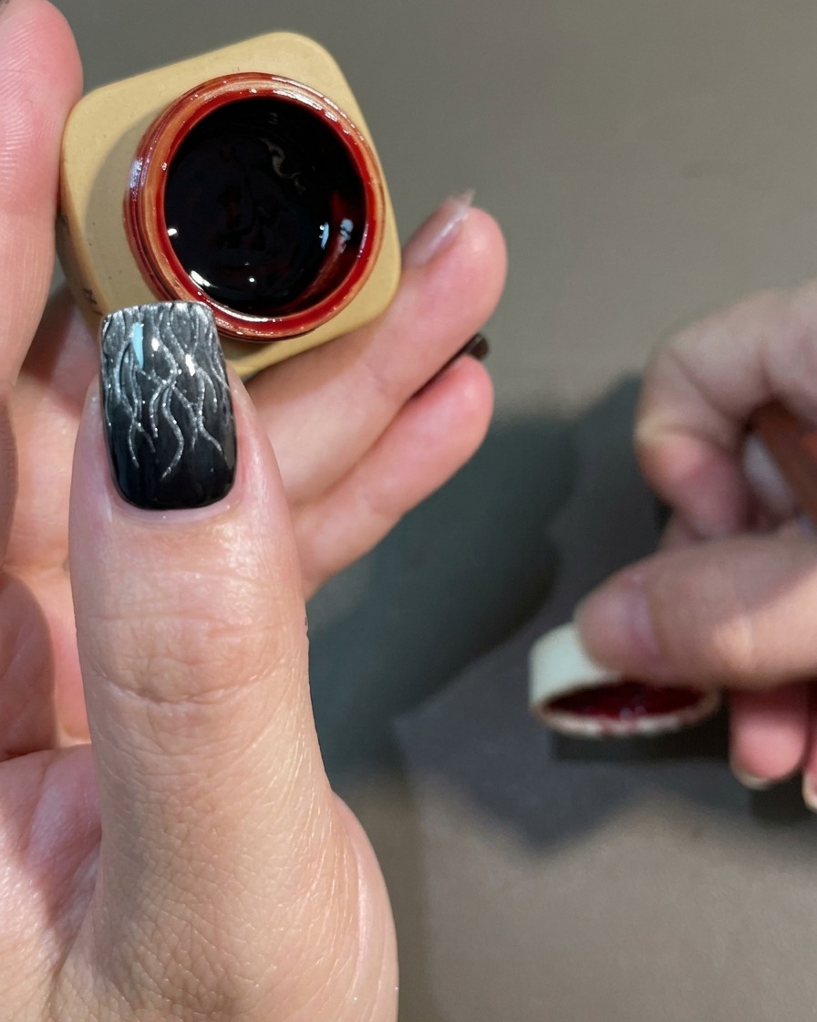 Un pennello per nail art che applica un gel rosso scuro semitrasparente su un design a fiamma cat-eye bianco e nero polimerizzato.