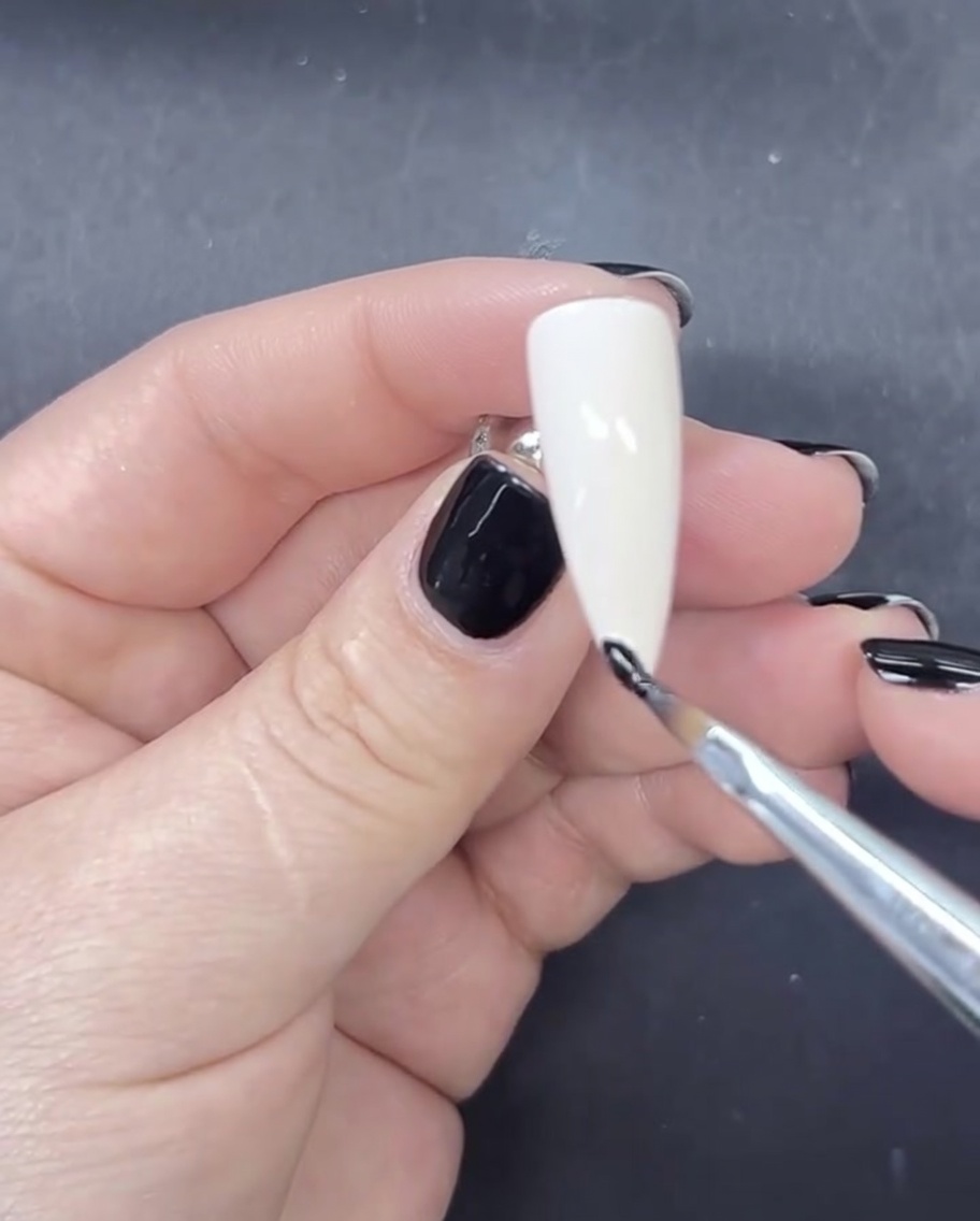 Un pennello liner sottile che applica una linea piccola e precisa di smalto gel nero sulla punta di ununghia dipinta di bianco latte.