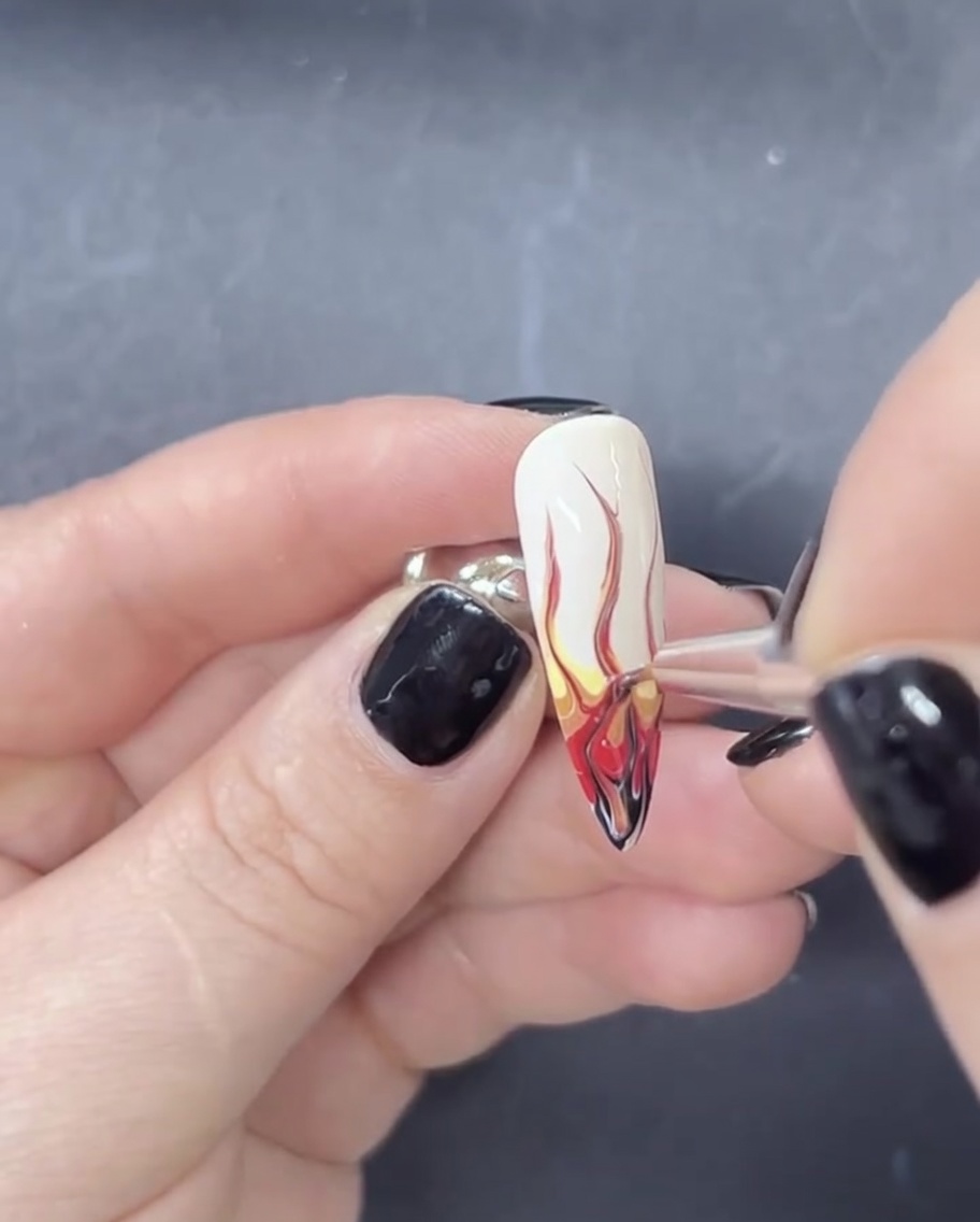 Un primo piano di un pennello liner per unghie che tira il gel rosso e nero verso lalto su una base bianca per creare forme di fiamma.