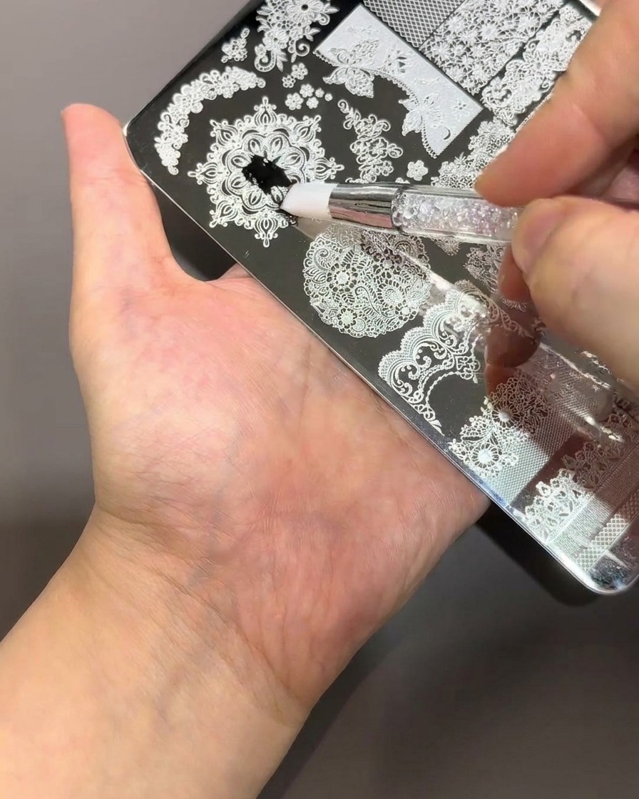 Un pennello applica smalto nero denso su una piastra per stamping in metallo argento con intricati motivi floreali e di pizzo.