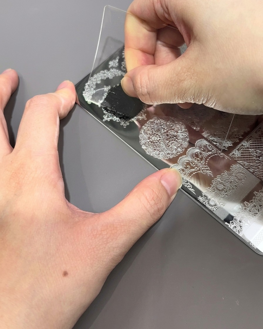 Una spatola in plastica trasparente viene trascinata su una piastra per stamping in metallo, spingendo via lo smalto nero in eccesso per rivelare il motivo inciso.