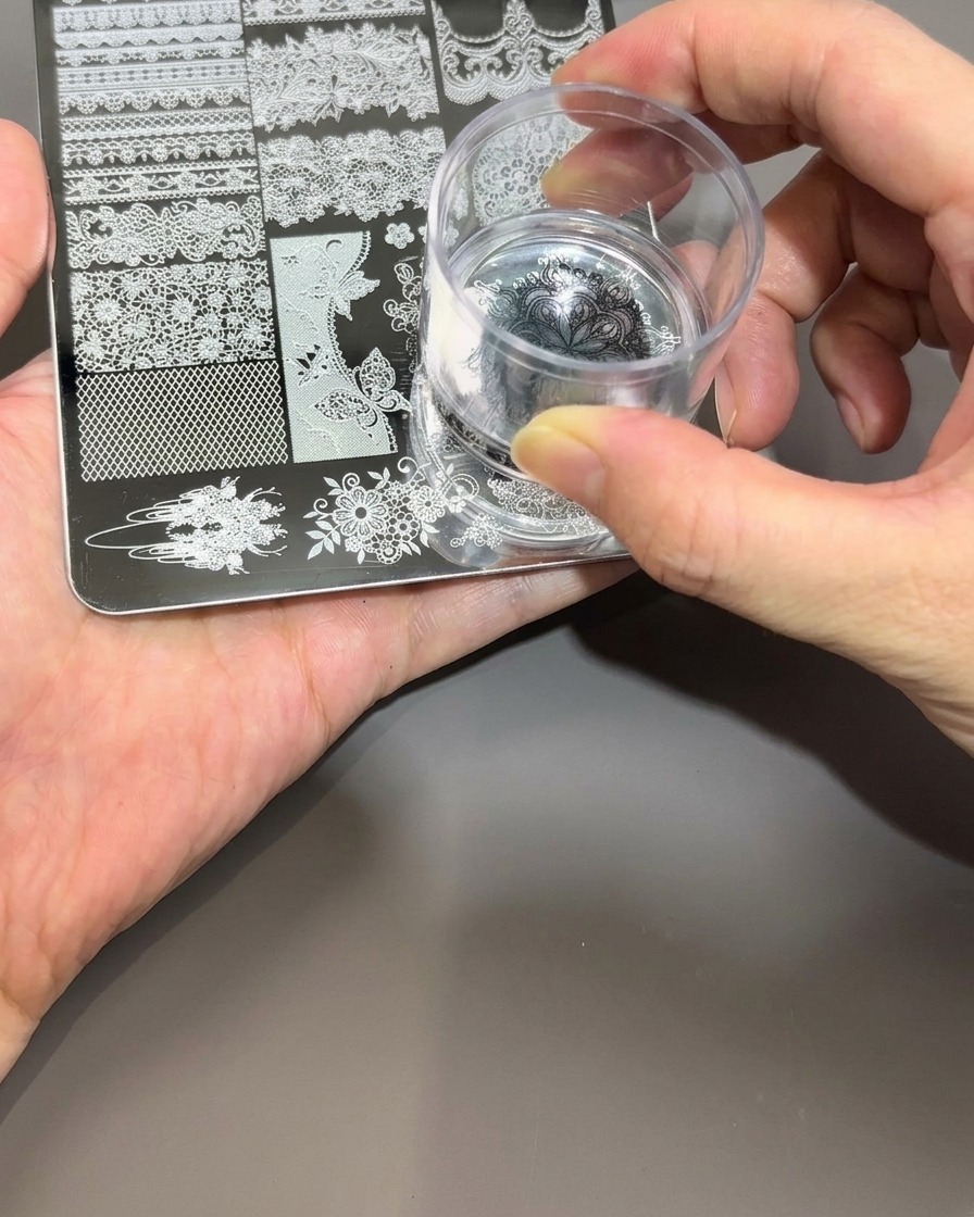 Una mano che tiene un timbro in silicone trasparente, premendolo delicatamente sul design in smalto nero su una piastra per stamping in metallo.
