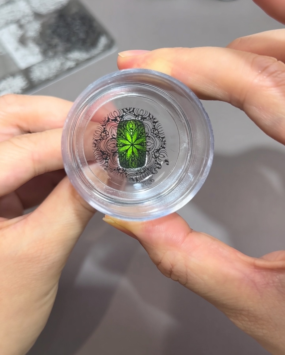 Vista attraverso un timbro in silicone trasparente, che mostra un intricato motivo nero perfettamente allineato su ununghia cat-eye verde brillante.