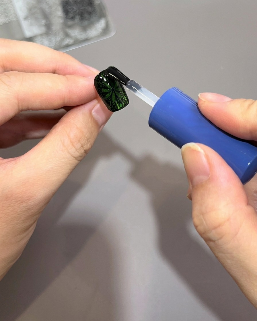 Un pennello per smalto applica uno strato spesso di top coat lucido trasparente su ununghia verde decorata con un intricato motivo stampato nero.