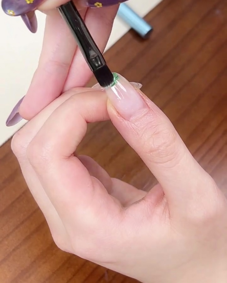 Un pennello piatto applica un piccolo punto di smalto gel verde chiaro sulla punta di ununghia naturale.