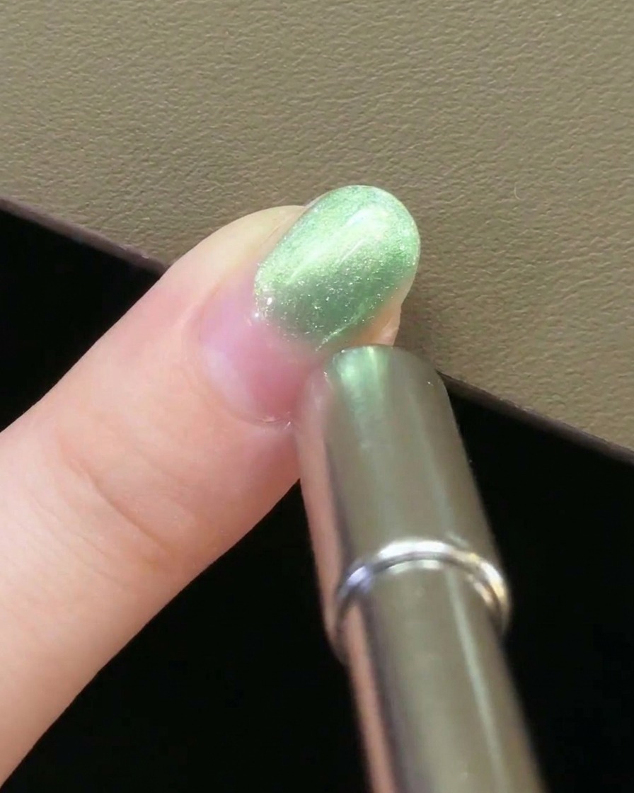 Un magnete per unghie in metallo tenuto parallelamente al lato di ununghia rivestita di gel magnetico verde.