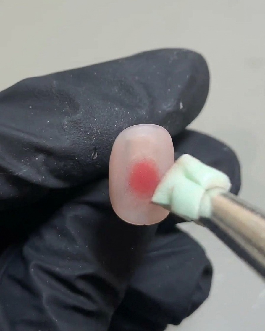 Pinzette che tengono una piccola spugna verde, picchiettando la goccia di gel rosa sullunghia.