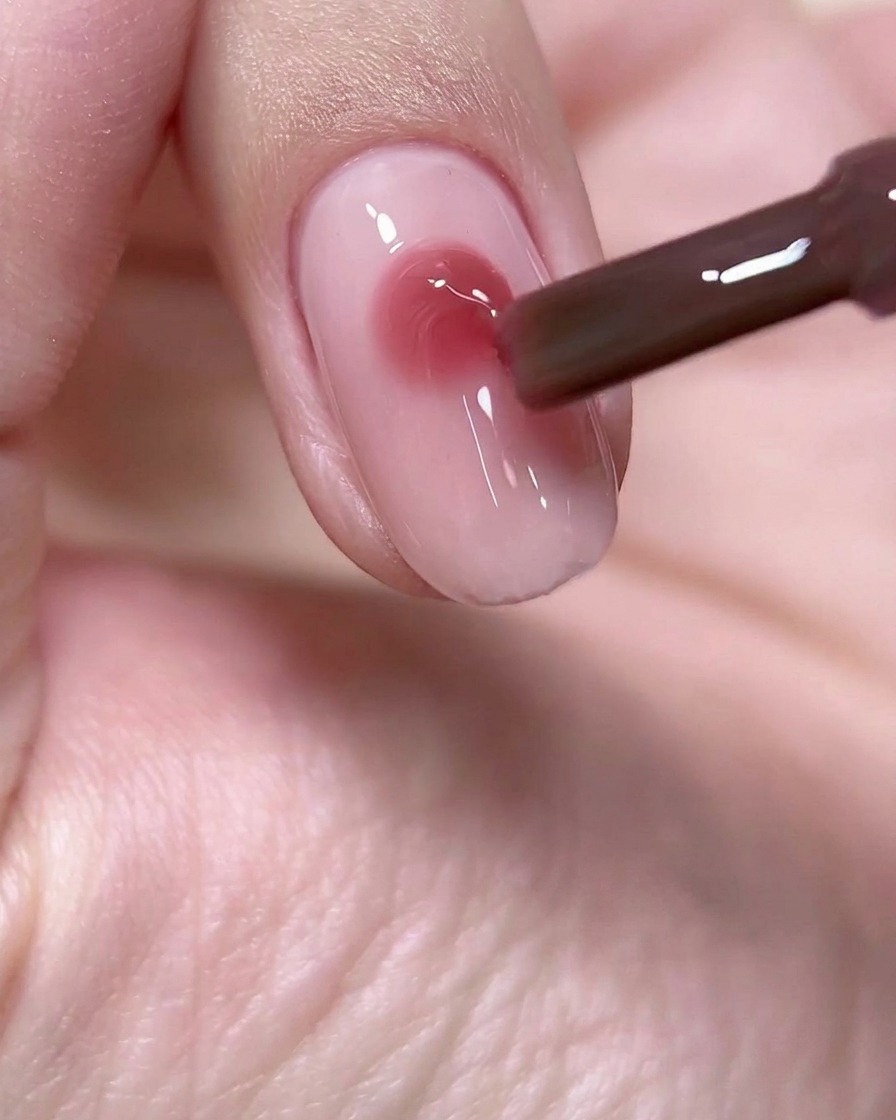 Un pennello da unghie che deposita una goccia precisa e rotonda di gel rosa vino al centro di una base per unghie rosa chiaro.