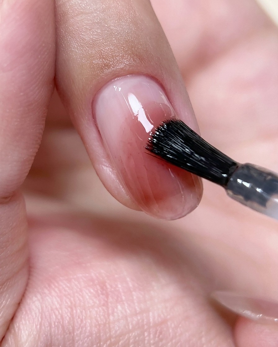 Un pennello da unghie che applica uno strato sottile e trasparente di top coat su una nail art blush rosa.