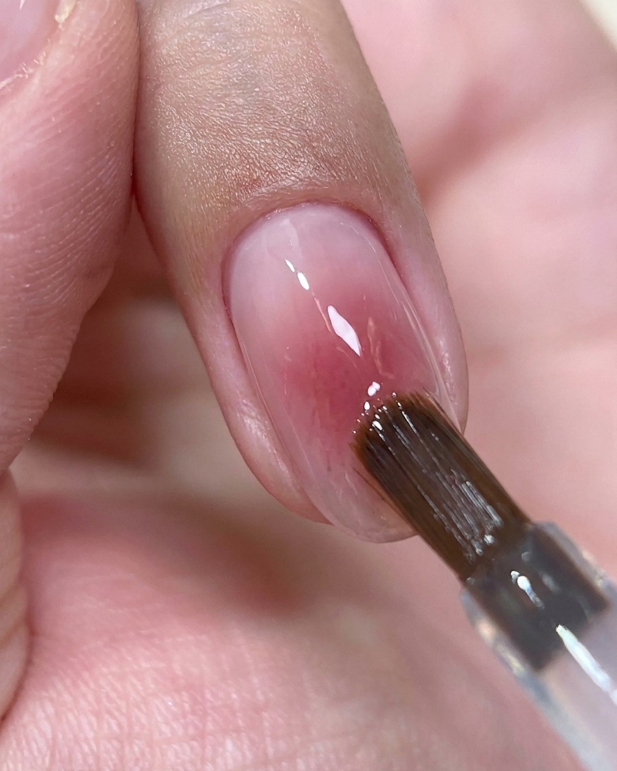 Un pennello da unghie che deposita una piccola goccia di top coat trasparente al centro di ununghia rosa lucida.