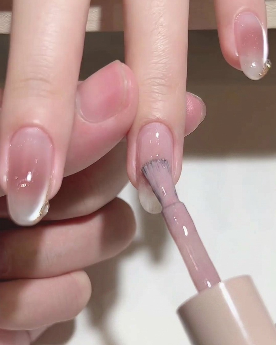 Primo piano di un pennello che applica uno smalto gel rosa traslucido su ununghia naturale.