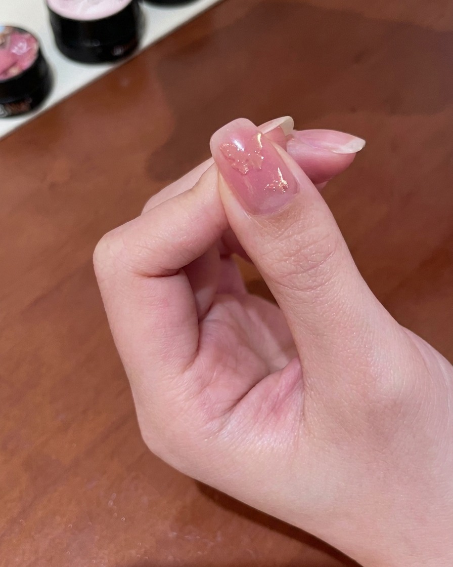 Primo piano di una mano che tiene un piccolo magnete cilindrico sopra ununghia rivestita di gel magnetico oro umido, rivelando uno shimmer mutevole.