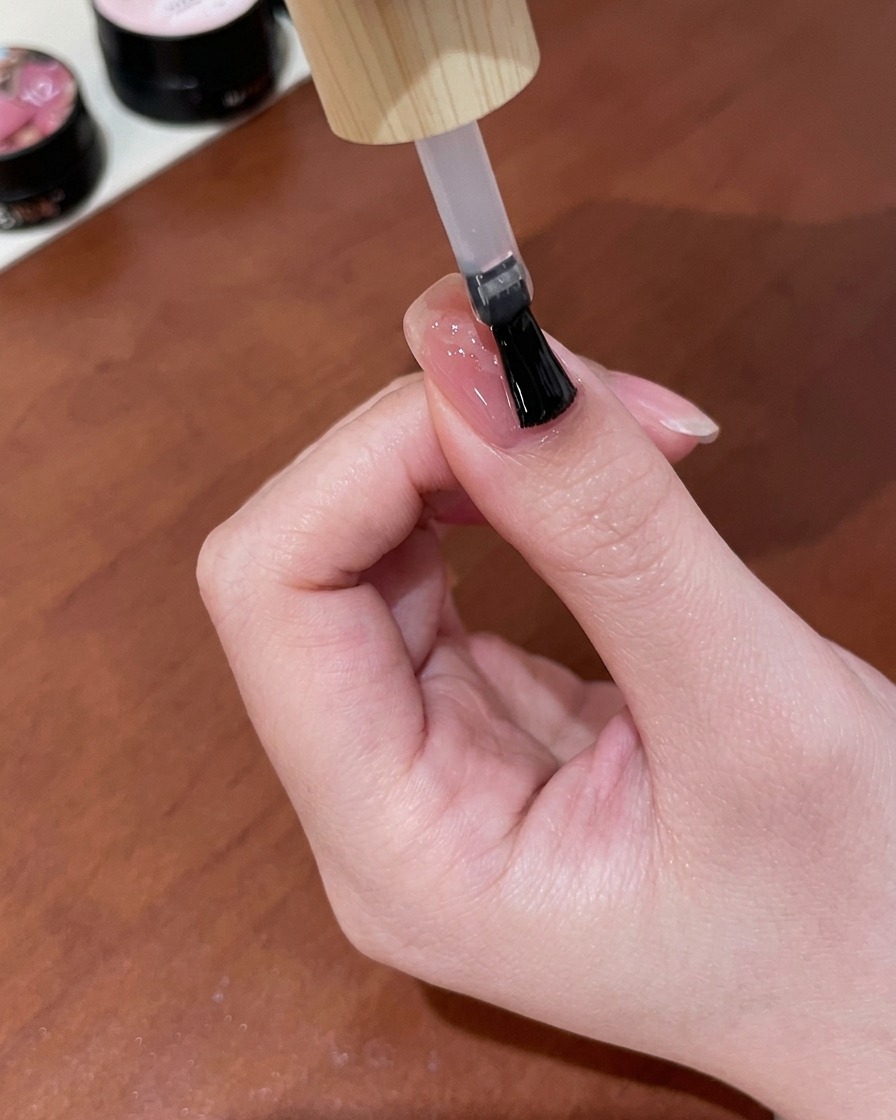 Un pennello che applica uno strato di top coat gel trasparente e brillante su ununghia nude pink finita con un design a farfalla cat-eye oro.