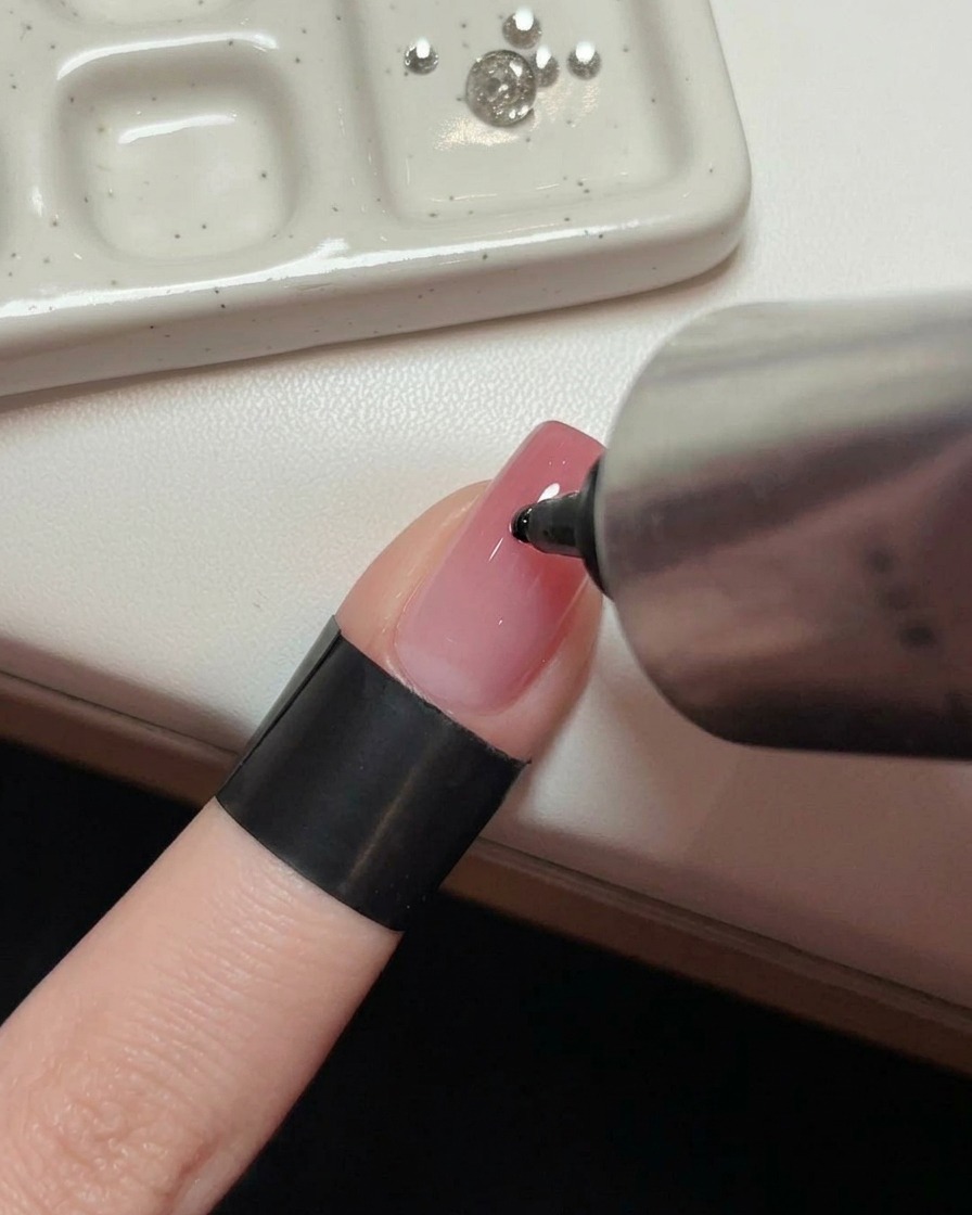 Una mano che tiene un tubetto di metallo mentre applica una goccia di gel adesivo trasparente al centro di ununghia rosa lucida.