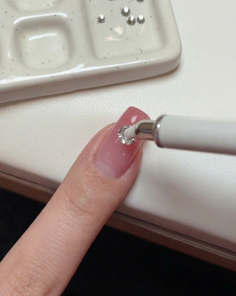 Uno strumento bianco con punta in cera che posiziona un grande cristallo rotondo da 5mm al centro di ununghia rosa.