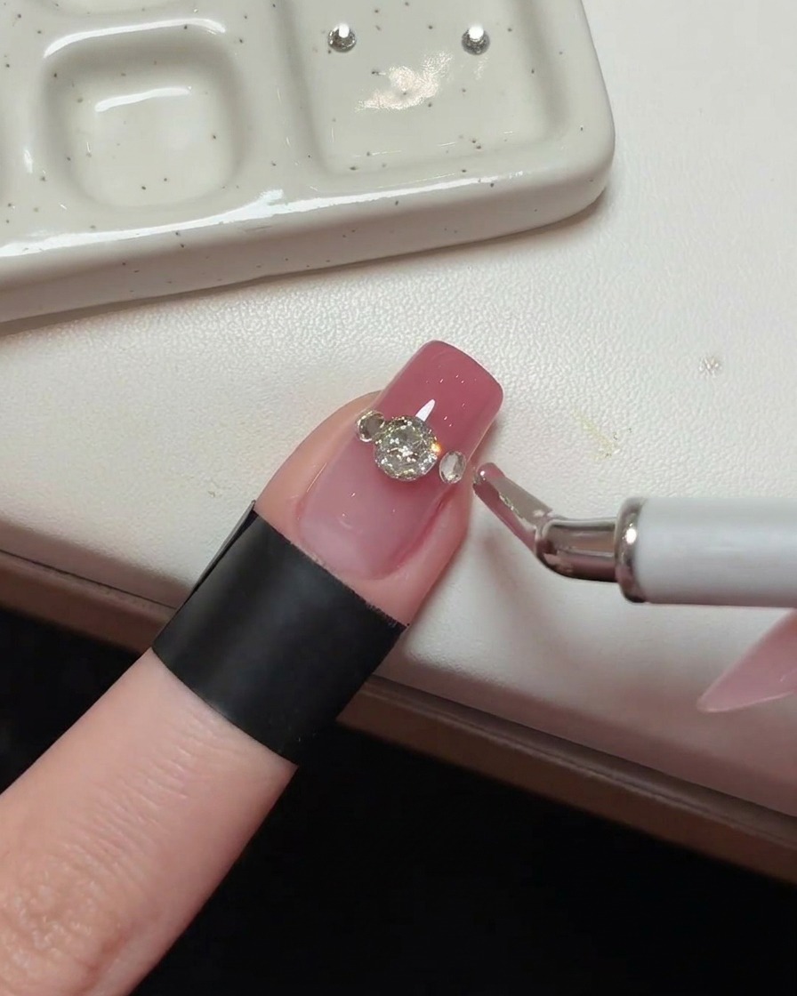 Primo piano di un cristallo principale su ununghia rosa, con cristalli decorativi posizionati ai lati.