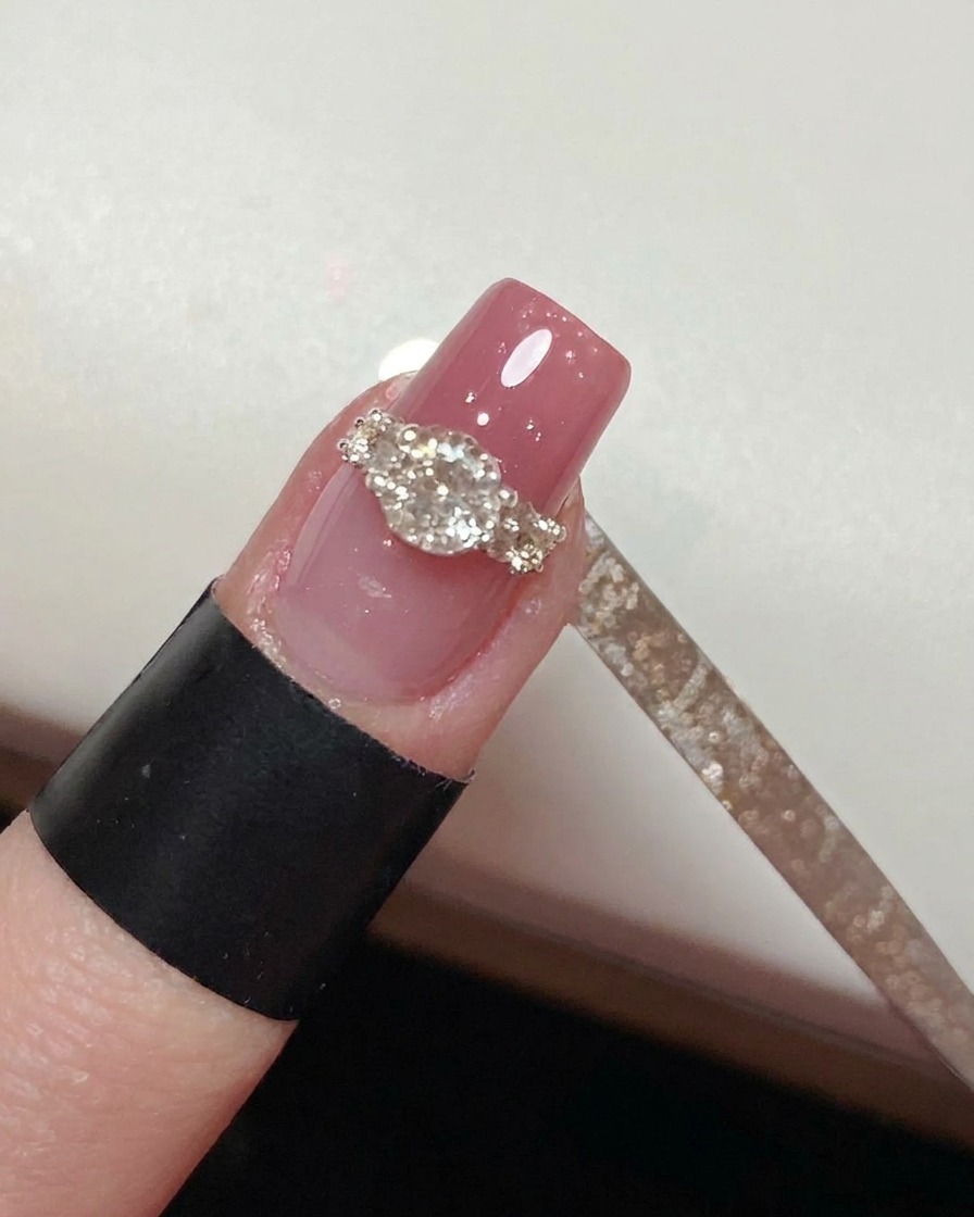 Un pennello a punta fine che dipinge accuratamente il top coat attorno alla base di un design ad anello con cristalli completato su ununghia rosa.