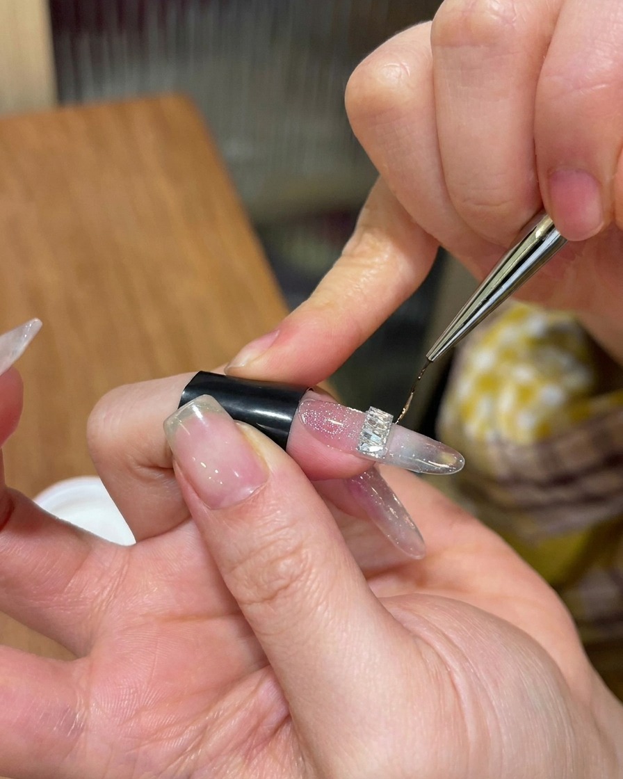Vista ravvicinata di un pennello da nail art sottile che applica gel top coat denso attorno alla base di uno strass argentato su ununghia glitterata.