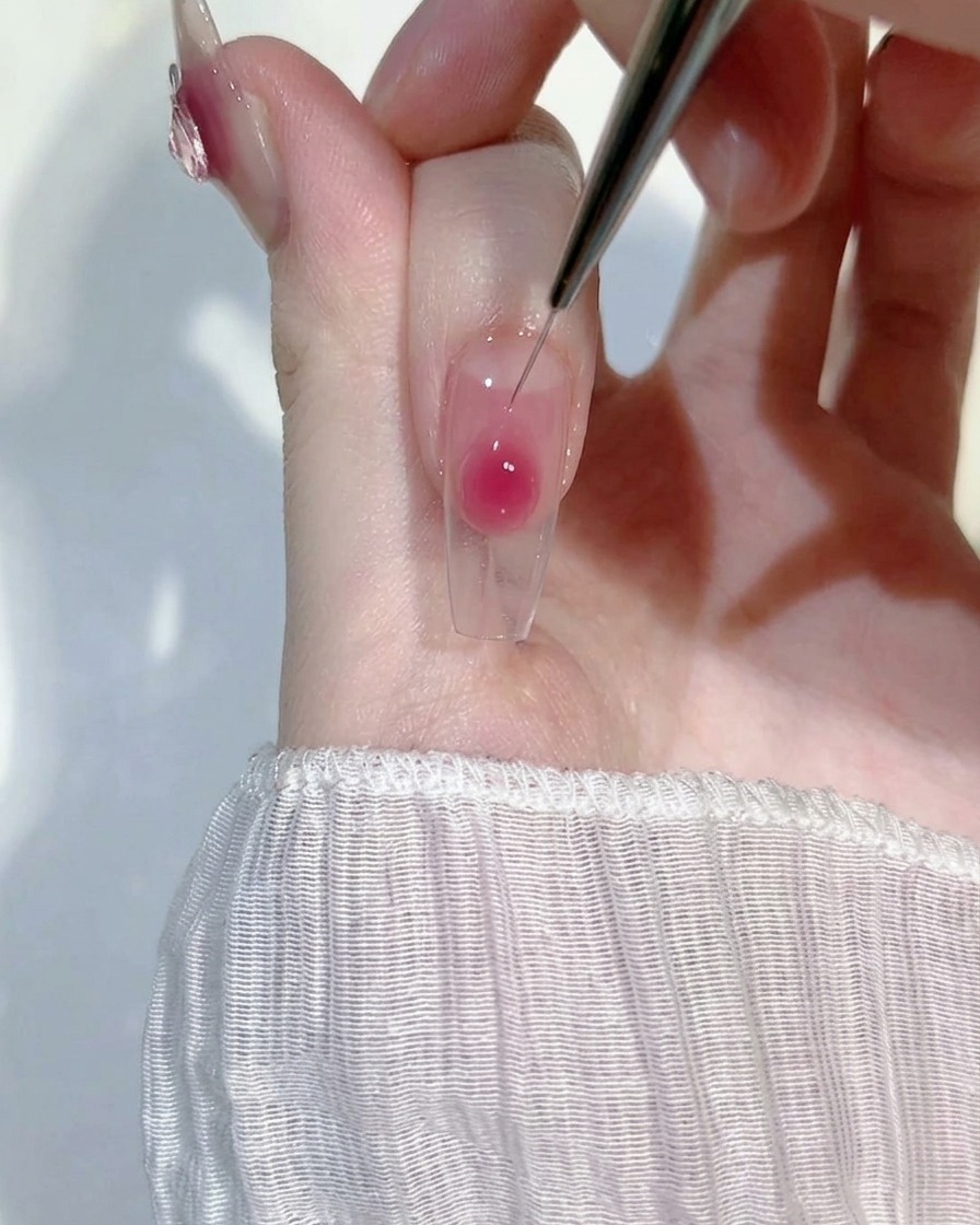 Primo piano di un pennello liner sottile che sfuma delicatamente una goccia di gel rosa su ununghia trasparente per un effetto gradiente.