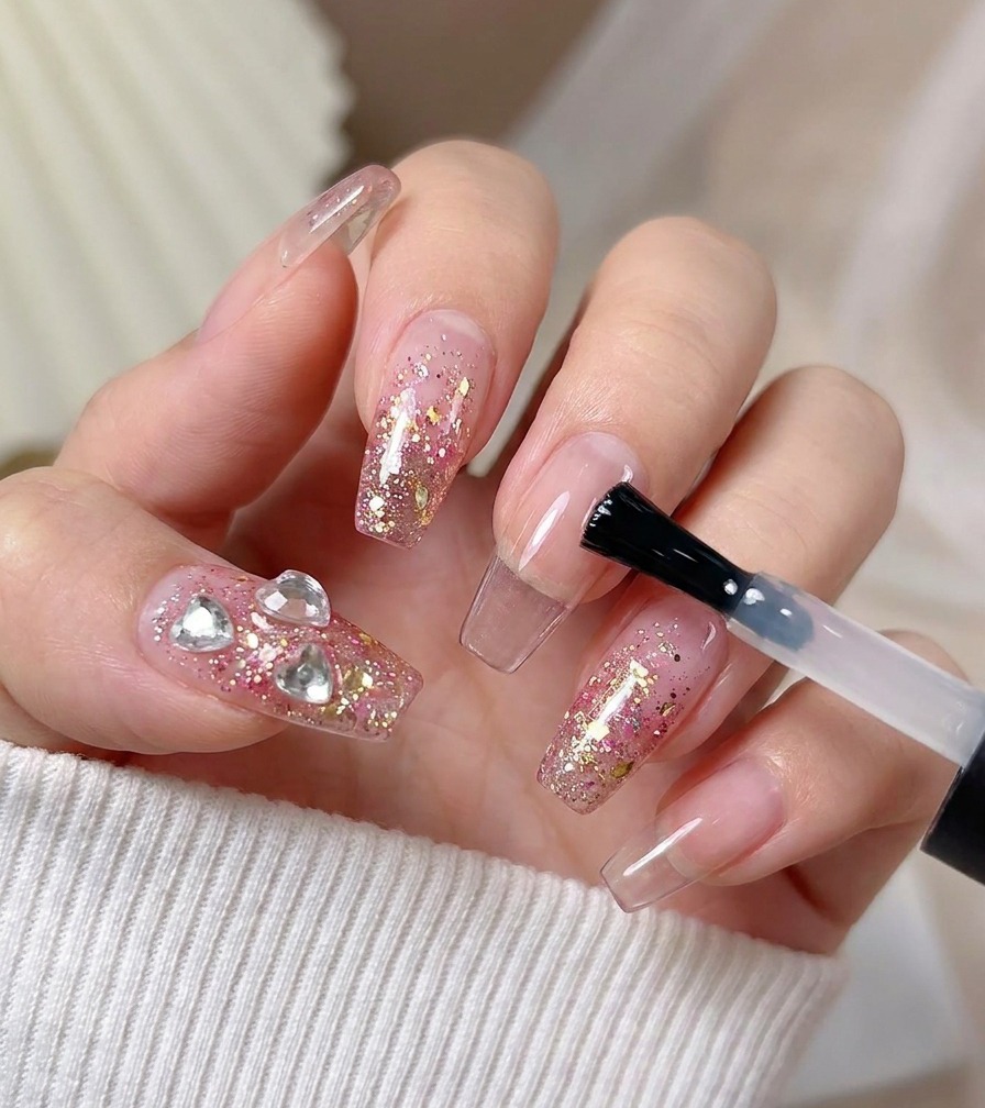 Un pennello applica un top coat lucido finale sopra ununghia con glitter rosa e cristalli a cuore.