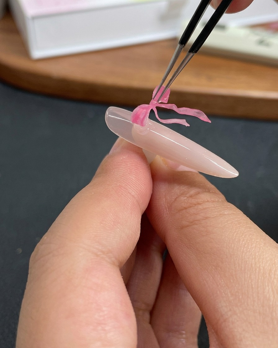 Pinzette che posizionano un delicato charm a fiocco a farfalla rosa su ununghia rivestita di gel trasparente.