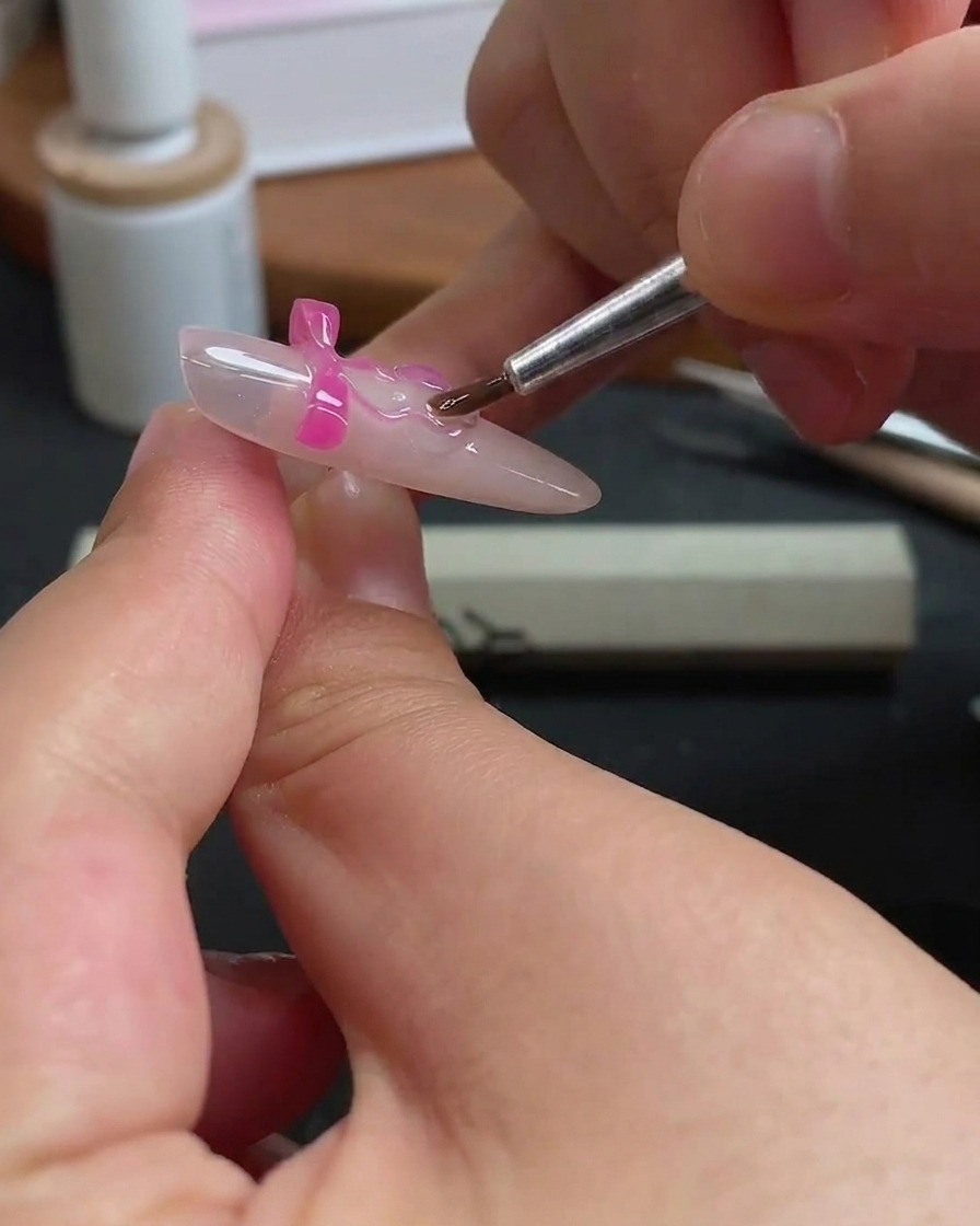 Un pennello sottile che applica con cura del gel trasparente attorno ai bordi di un fiocco rosa su ununghia.