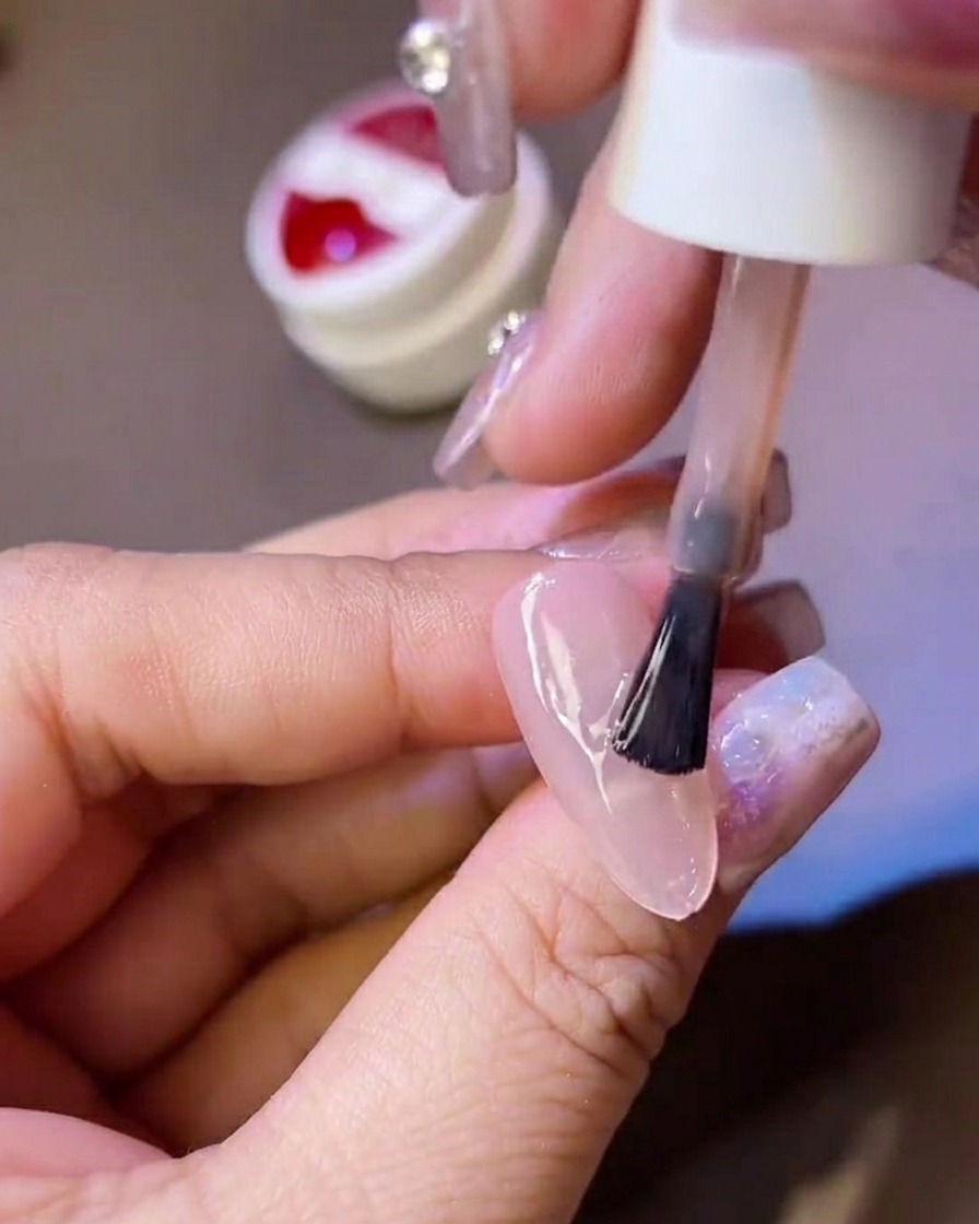 Una mano che tiene un pennellino, applicando uno strato di top coat opaco su una tip per unghie traslucida.