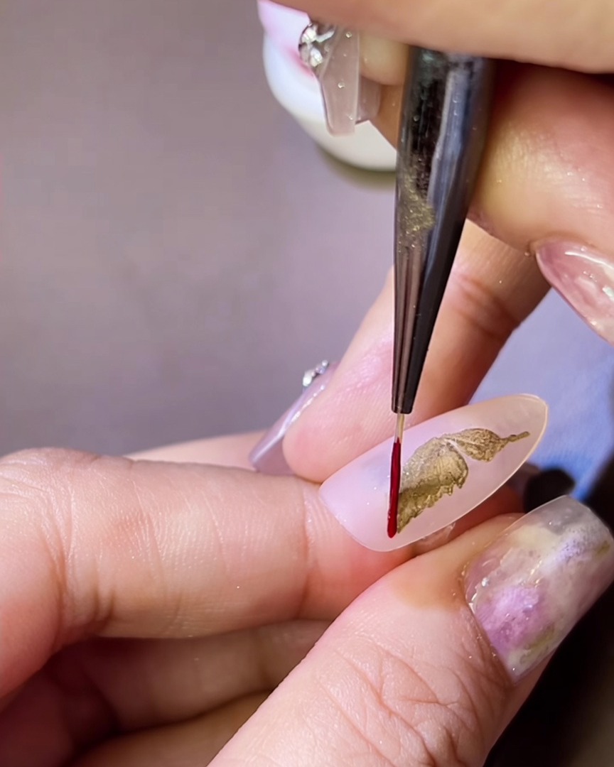 Un pennello a punta fine che applica un sottile contorno in gel rosso attorno a unala di farfalla metallica dorata su ununghia.