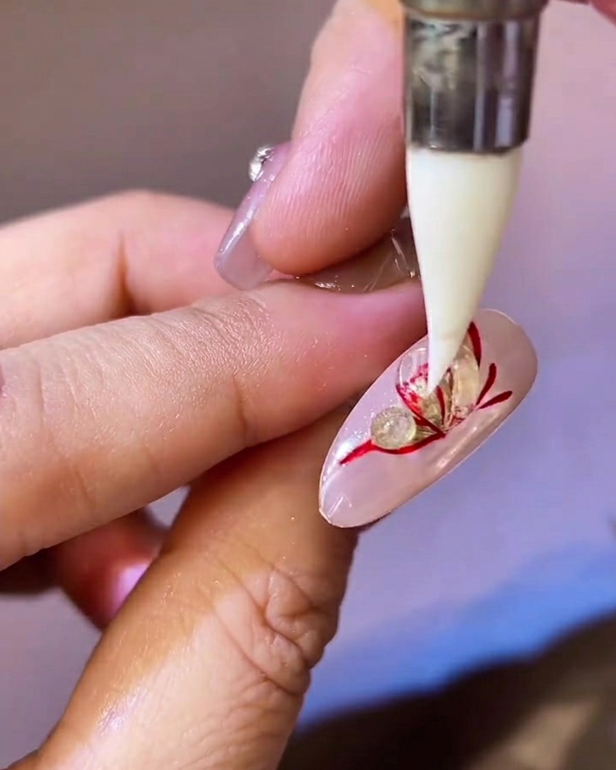 Uno strumento in silicone a punta che preme e appiattisce una perla di gel scultoreo trasparente su unala di farfalla rossa e oro su ununghia.