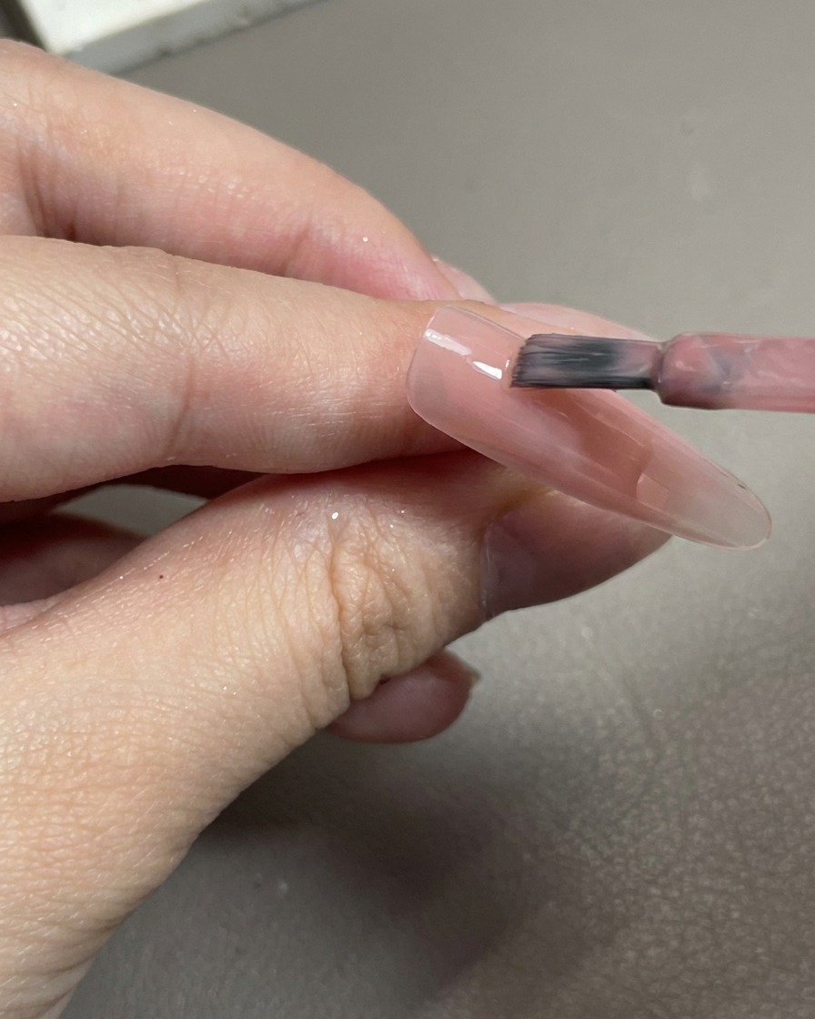 Primo piano di un pennello che applica smalto gel rosa traslucido su una punta di unghia trasparente.