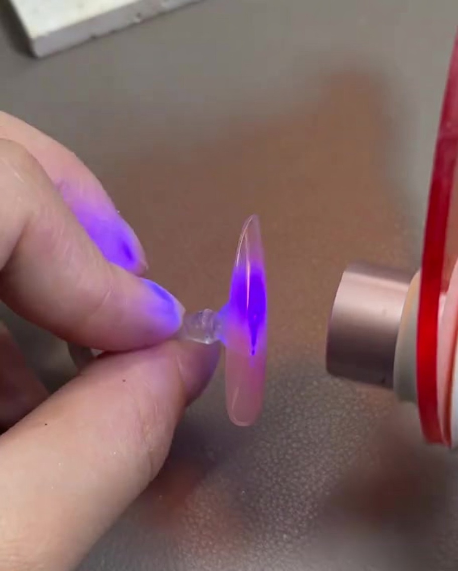 Una mano che tiene una punta di unghia trasparente con gel rosa sotto una mini lampada UV/LED per unghie.