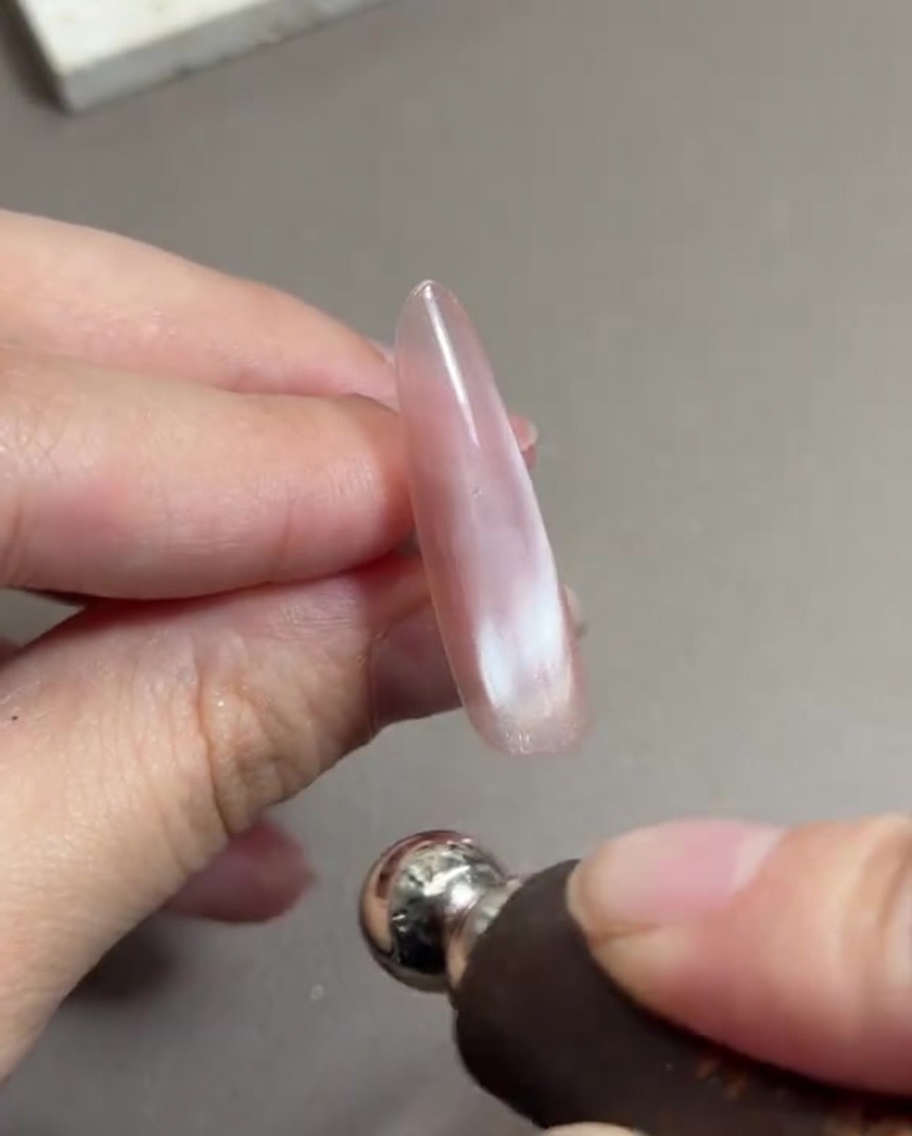 Una mano che tiene uno strumento magnetico cilindrico vicino a una lunga unghia dipinta con gel cat-eye rosa bagnato, spostando le particelle magnetiche.